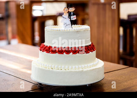 Weisse Hochzeitstorte mit Braut und Bräutigam Figuren an der Spitze. Schöne weisse Hochzeitstorte mit roten Rosen in der Tabelle mit den kleinen Mann und Frau Marionetten Stockfoto