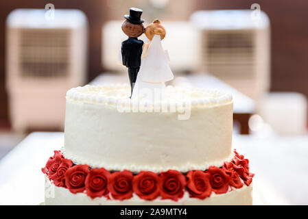 Weisse Hochzeitstorte mit Braut und Bräutigam Figuren an der Spitze. Schöne weisse Hochzeitstorte mit roten Rosen in der Tabelle mit den kleinen Mann und Frau Marionetten Stockfoto