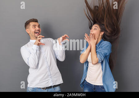 Spaß haben. Junges Paar Mann erschreckend Frau verspielt Studio stehen isoliert auf Grau Stockfoto