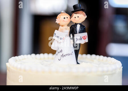 Weisse Hochzeitstorte mit Braut und Bräutigam Figuren an der Spitze. Schöne weisse Hochzeitstorte mit roten Rosen in der Tabelle mit den kleinen Mann und Frau Marionetten Stockfoto