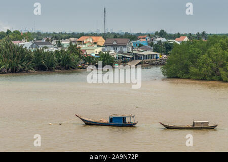 Lange Tau Fluss, Vietnam - März 12, 2019: 2 schaluppen sind auf braunem Wasser verankert. Phuoc Khanh Riverside Village mit mehreren Häusern. Licht blauer Himmel und Stockfoto