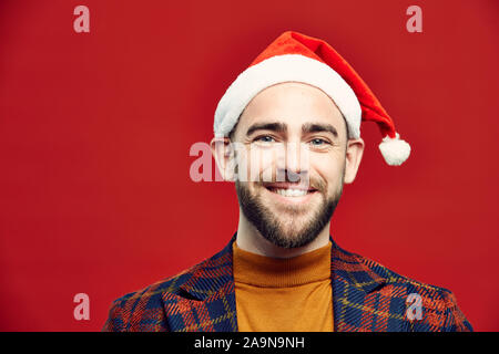 Kopf und Schultern Portrait von bärtigen Mann mit Weihnachtsmütze fröhlich in die Kamera lächelt, während gegen den roten Hintergrund posiert, Kopie Raum Stockfoto