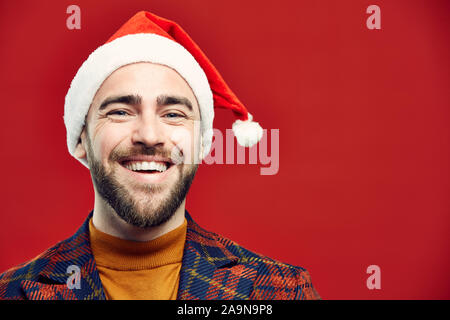 Kopf und Schultern Porträt des modernen erwachsener Mann, Santa hat freundlich in die Kamera lächelnd, während gegen den roten Hintergrund posiert, Kopie Raum Stockfoto