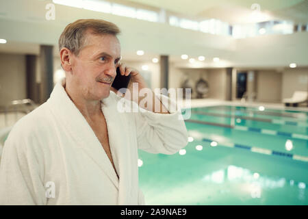 Taille bis Porträt des modernen älterer Mann, Bademantel und Sprechen telefonisch beim Stehen mit Schwimmbad im luxuriösen Spa, Kopie Raum Stockfoto