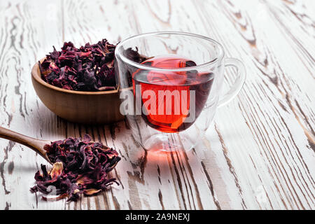 Red Hibiscus Tee im Glas Schale auf weiße Holztisch mit trockenen Rosenblätter, Löffel und Schüssel Stockfoto