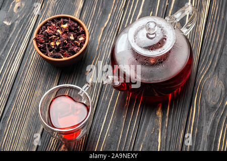 Blick von oben auf die roten Hibiskus Tee Teekanne aus Glas und Herzförmige Schale auf Holztisch mit trockenen Rosenblüten in der Schüssel Stockfoto