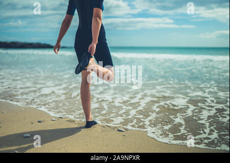 Eine junge Frau in einem Neoprenanzug am Strand setzt sich auf Ihre wasserdichte Schuhe Stockfoto