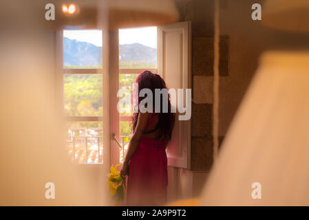 Frau, die durch ein Fenster mit Blick auf die Berge, in einem rosa Kleid mit gelben Blumen in der Hand. Stockfoto