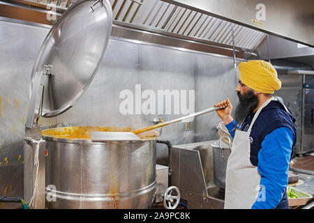 Ein Sikh männliche Freiwillige Köche Tofu in einem sehr großen Topf. In einem Langar, eine kostenlose Gemeinschaftsküche. In South Richmond Hill, Queens, New York City. Stockfoto