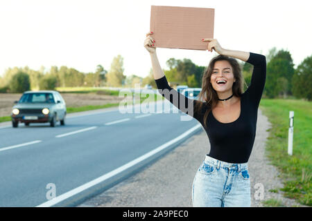 Junge Frau per Anhalter auf der Straße ist mit einem leeren Karton Zeichen Stockfoto