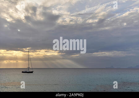 Segelschiff bei Sonnenuntergang mit Wolken und Sonnenstrahlen in der Nähe von Cala Saona mit Es Vedrá und Ibiza Inseln in der Ferne (Formentera, Balearen, Spanien) Stockfoto