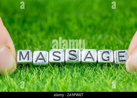 Nahaufnahme auf Wort MASSAGE in Metall Buchstaben auf Gras gelegt geschrieben und zwischen die Finger einer Frau. Konzept von Wellness Hintergrund Stockfoto