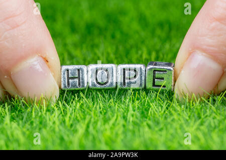 Nahaufnahme auf Wort Hoffnung in Metall Buchstaben auf Gras gelegt geschrieben und zwischen die Finger einer Frau. Konzept der Sehnsucht, Hoffnung Hintergrund Stockfoto