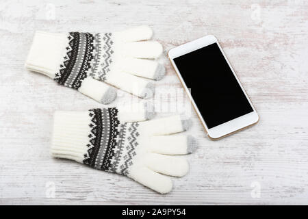 Winter Handschuhe aus Gewirken und smart phone mit leeren Bildschirm auf weißem Holz- Hintergrund. Smartphone Mock-up mit leeren schwarzen Bildschirm. Stockfoto