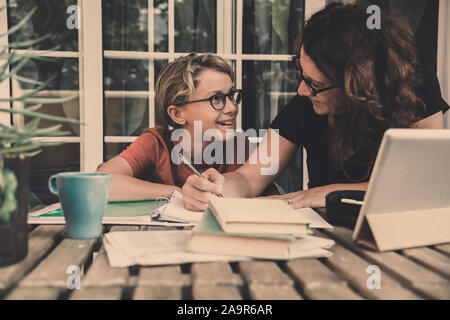 Junge Schüler Hausaufgaben zu Hause mit Schule Bücher, Zeitung und digitale Pad von seiner Mutter geholfen. Mom schreiben auf dem copybook Lehre seinen Sohn. Stockfoto