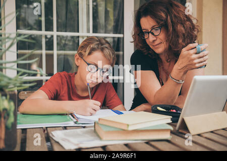 Junge Schüler Hausaufgaben zu Hause mit Schule Bücher Zeitung, Digital Pad von seiner Mutter geholfen. Mom control, Hilfe und Lehre seinen Sohn. Bildung, Stockfoto