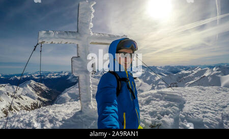 Ein Mann in Ski Outfit stehen auf der Spitze eines Berges mit einem schneebedeckten Kreuz hinter ihm. Endlose Schnee caped Berge rund um den Mann. Weich Stockfoto