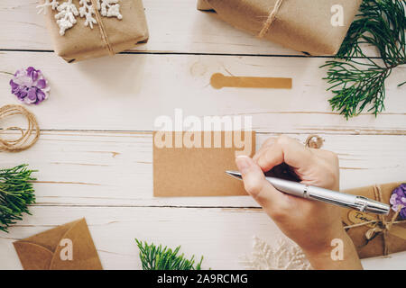 Nahaufnahme von Händen Frau schreiben leer Merkzettel und Weihnachten Karte auf Holztisch mit Weihnachten Dekoration. Stockfoto