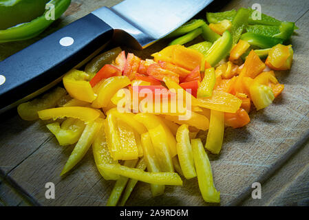 In Scheiben geschnitten bunt rot, gelb, grün und orange Klingel oder Paprika auf alten zerkratzte Holz Schneidebrett verwendet. Großes Metall Messer ist in der Nähe des pep Stockfoto
