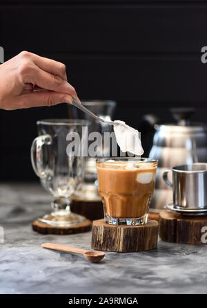 Frau Hand gießen Schlagsahne Kokoscreme in Blended vietnamesischen Filterkaffee in Glas auf dunklem Hintergrund Kopie Raum selektiven Fokus Stockfoto