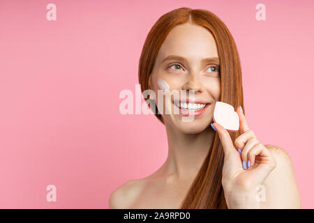 Headshot schöne fröhliche rothaarige Frau mit Sauber Gesunde Haut, das Anwenden der Grundlage mit kosmetischen Schwamm, lächelnd weiße Zähne auf isolierte Stockfoto