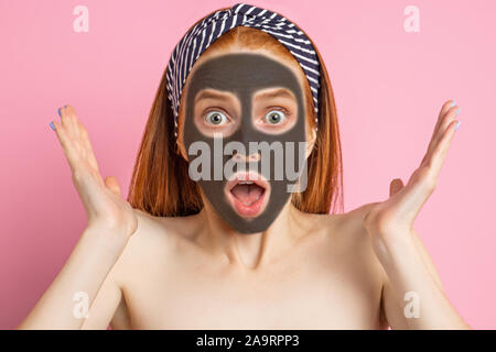 Nahaufnahme von erstaunt verwirrten jungen kaukasischen Frau mit Maske im Gesicht, an der Kamera mit schockiert Ausdrücken suchen, tragen Stirnband, Posieren auf Rosa Stockfoto