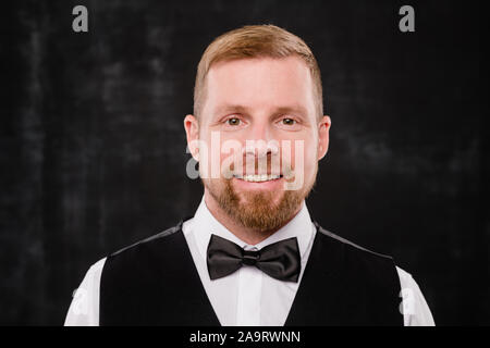 Glückliche junge elegante Kellner in schwarze Weste und Fliege zu Ihnen schauen Stockfoto