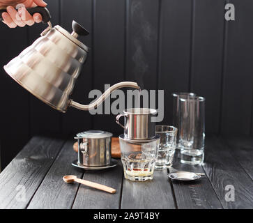 Preraring Filterkaffee vietnamesischen Stil. Frau Hand gießen dampfend heißes Wasser aus Schwanenhals Wasserkocher in Metall/Teekocher phin auf schwarzem Hintergrund Stockfoto