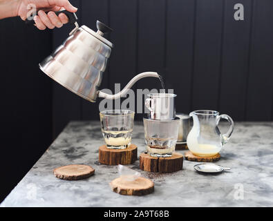 Die vietnamesischen Kaffee Milch darübergießen. Frau Hand gießt Wasser von Schwanenhals Wasserkocher in phin auf dunklem Hintergrund Kopie Raum Stockfoto