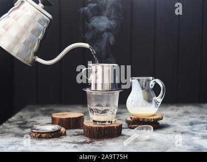 Die vietnamesischen Kaffee mit Kondensmilch. Strömenden heißen Wasser aus Schwanenhals Wasserkocher in Glas durch phin auf dunklem Hintergrund Kopie Raum Stockfoto