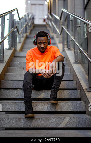 Junge afrikanische amerikanische Mann im stilvollen orange Hoodie Sweatshirt in drahtlosen Kopfhörern Musik hören und sitzen auf der Treppe. Stockfoto
