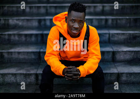 African-american man im stilvollen orange Hoodie Sweatshirt sitzen auf Treppen Stockfoto
