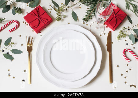 Weihnachten Tisch setzen. Golden Besteck, Porzellan Teller, Champagner Glas, Tanne, Eukalyptus Zweige, rot Geschenkboxen und golden Konfetti Stockfoto