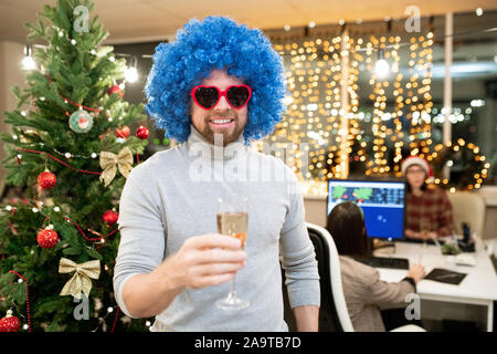 Fröhlicher junger Geschäftsmann, der in blauen Lockenperücke Toasten mit Flöte Champagner Stockfoto