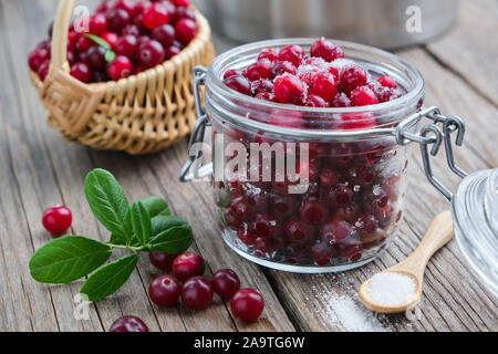 Jar reife Preiselbeeren mit Zucker, Korb von bog Beeren und Kochtopf auf Hintergrund. Stockfoto