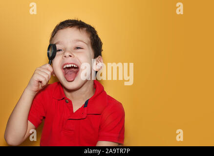 Ein kleiner Junge schaut durch eine Lupe in rotem T-Shirt, auf purem gelben Hintergrund. Junge lacht und zeigt Zunge. Leerzeichen für Text Stockfoto