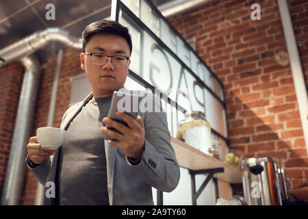 Low Angle Portrait von younf asiatischer Mann holding Smartphone und Tasse Kaffee bei der Arbeit im Büro, Kopie Raum Stockfoto