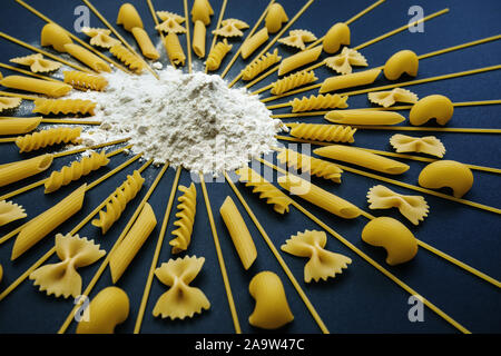 Pasta angeordnet um Stapel von Mehl und Weizen. Geometrische Anordnung unterschiedlicher Art der Nudeln auf schwarzem Hintergrund. Stockfoto