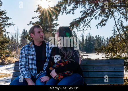 Ein Paar in den 30er Jahren, mit einem weißen Chihuahua, sitzen auf einer Parkbank, in einem besonderen Moment, umgeben von Bäumen und einem hellen Sunburst umgeben. Stockfoto