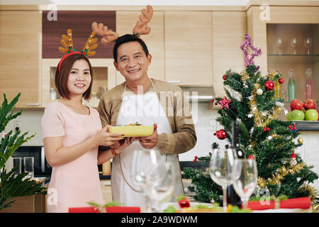 Glücklich lächelnde reife asiatische Paar tragen Rentier Geweih beim Servieren Tisch für das Weihnachtsessen Stockfoto