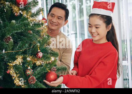 Recht jugendlich asiatischen Mädchen im roten Pullover schmücken Weihnachtsbaum mit ihrem Vater. Stockfoto