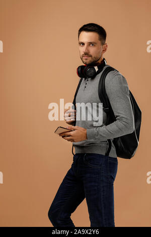 Hübscher junger Mann mit Kopfhörer und Rucksack mit Smartphone auf beigen Hintergrund isoliert. Stockfoto