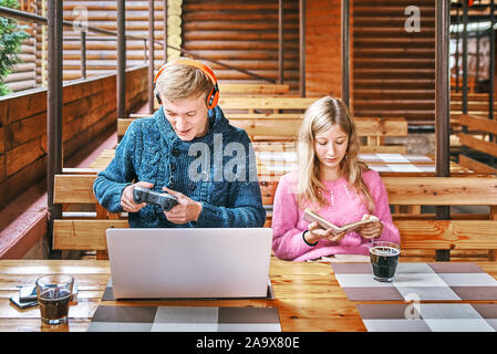 Jungen Kerl spielt Spiele bei einem Laptop in einem Cafe. das Mädchen in der Nähe ist ein Buch zu lesen. Ein Verschleiß Konzept Stockfoto