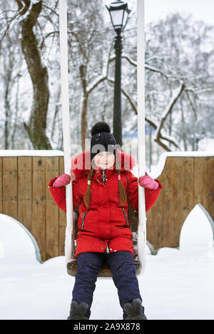 Winter Portrait eines kleinen Mädchens in einen Hut und eine rote Jacke auf einer Schaukel Stockfoto