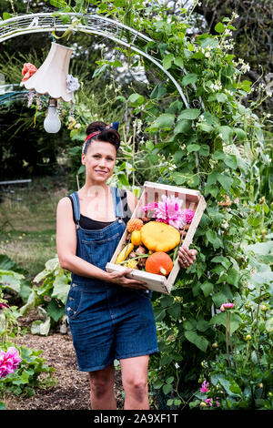 Frau, die in einem Garten, der Holzkiste mit frischem Gemüse und Blumen, in die Kamera lächeln. Stockfoto