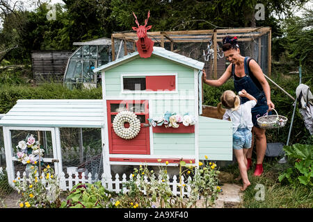 Lächelnde Frau und Mädchen stehen in einem Garten neben einem bunten Hühnerstall. Stockfoto