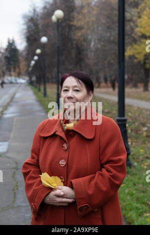 Eine ältere Frau in einem hellen Terrakotta Fell sitzt auf einer Bank in einem Park im Herbst mit einem Laptop und wählt Waren online Stockfoto