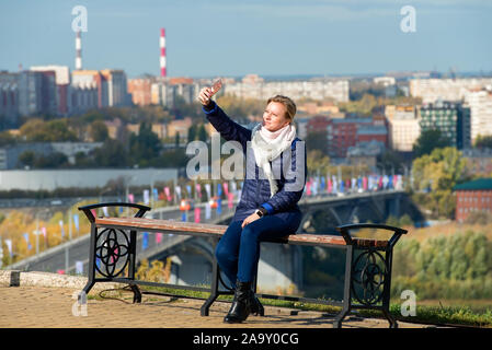 Nischni Nowgorod, ein Mädchen sitzt auf einer Bank und nimmt eine selfie vor dem Hintergrund der Stadt unten, im sonnigen Herbsttag Stockfoto