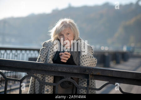 Im Alter von Obdachlosen Frau mit Mantel auf Schultern holding Tasse Tee Stockfoto