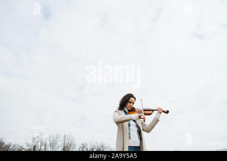 Schöne Frau in einen Mantel spielen Geige unter einem Himmel mit geschlossenen Augen Stockfoto